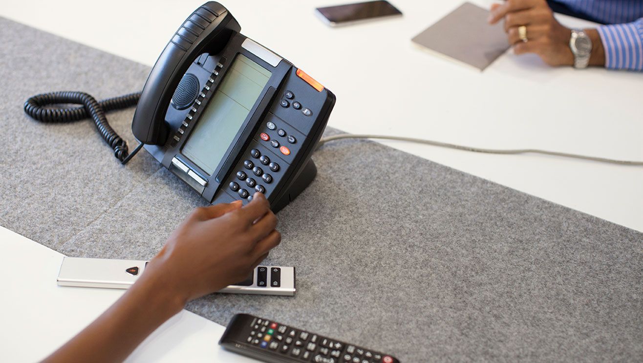 Two people sit facing each other with a desk phone and two remote controls in the centre of the desk