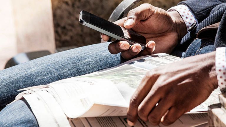 A person sitting with a newspaper on their lap looks at their smartphone