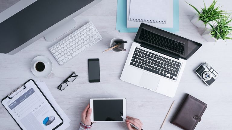 A desk arranged with a desktop computer, laptop, smart phone and camera