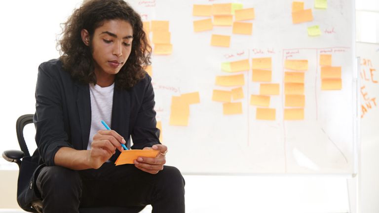 A woman holds a pad of sticky notes in front of a board covered in sticky notes
