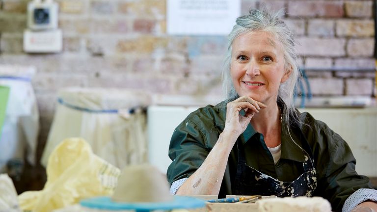 A woman wearing overalls sits behind a piece of clay on a table
