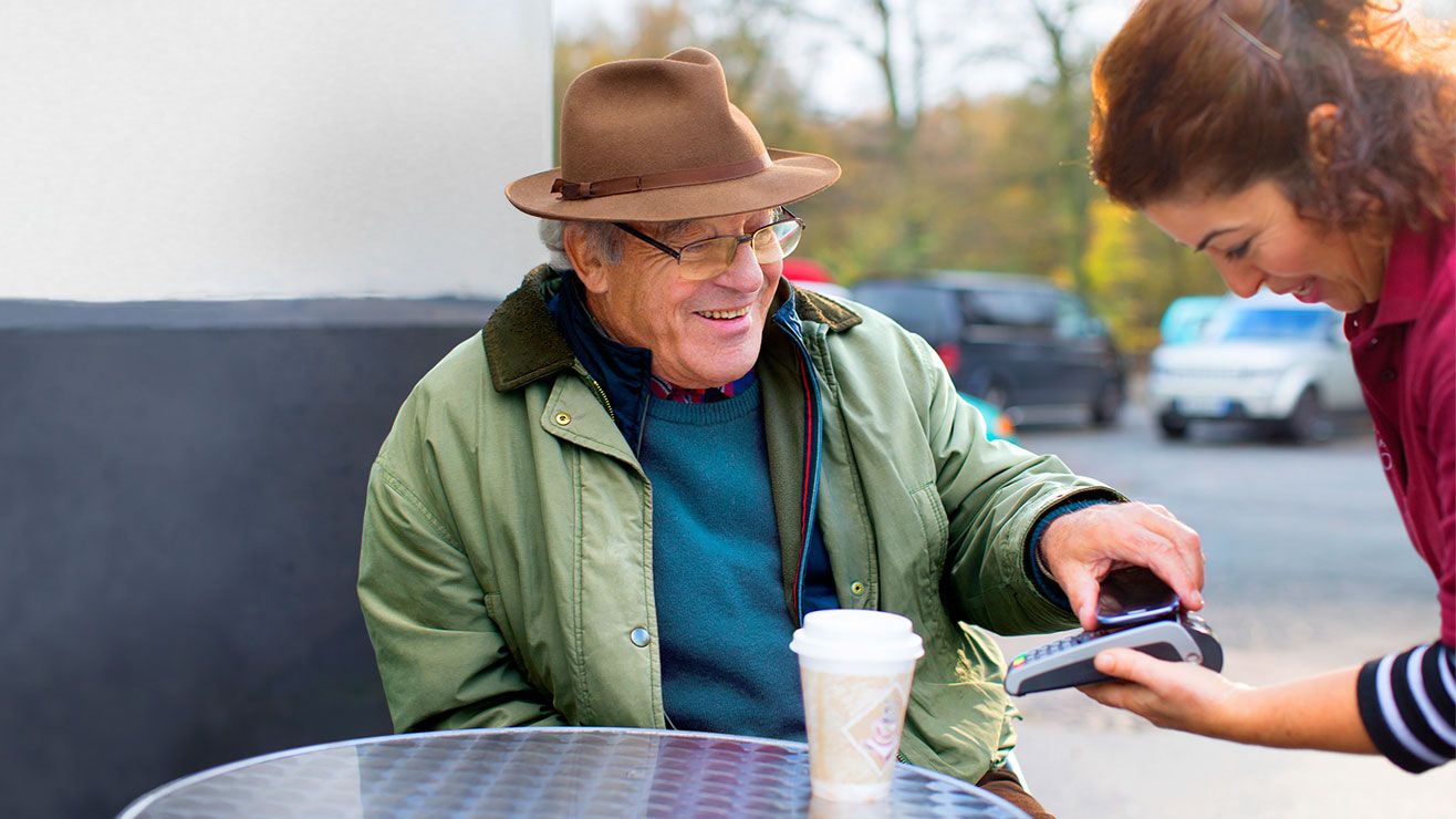 A man sitting at an outside table with a coffee, taps his card on a card reader held by a waitress
