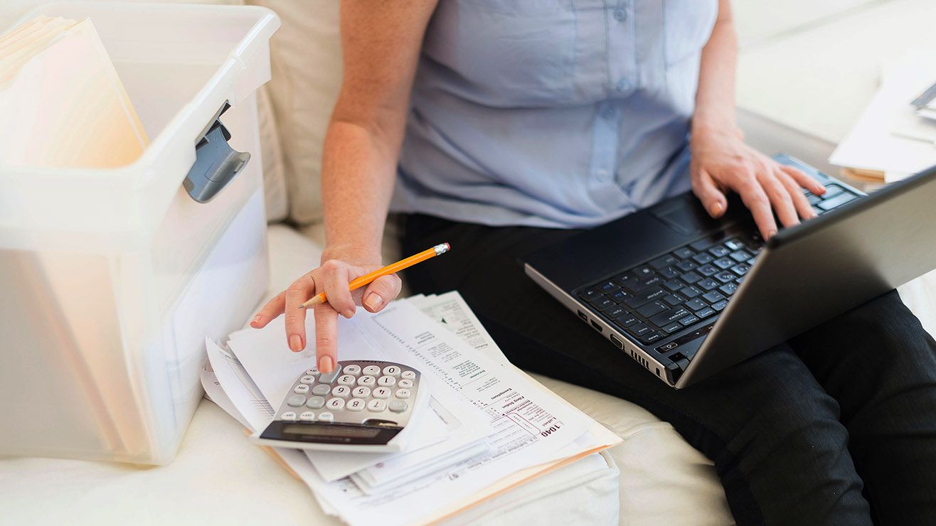 A person seated with a laptop on their lap taps the keys with one hand and a calculator with the other
