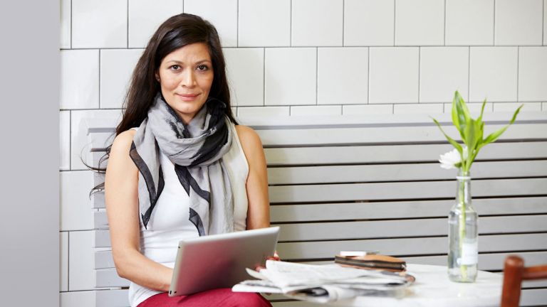 A woman holding a tablet sits in a restaurant with a newspaper on the table