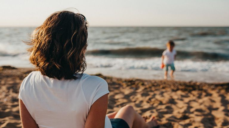  A woman sits on a sandy beach watching a child playing in the surf